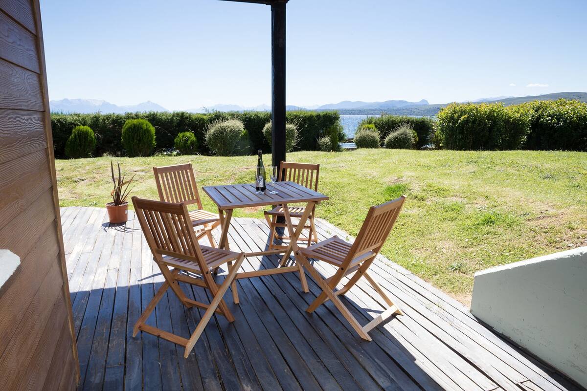 Terraza de madera con una mesa y sillas, con vistas a un área verde y montañas junto a un cuerpo de agua.