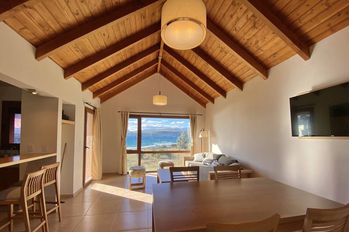 Amplia habitación con techo de madera alto, un comedor con mesa y sillas, un sofá, y un gran ventanal con vistas al mar.