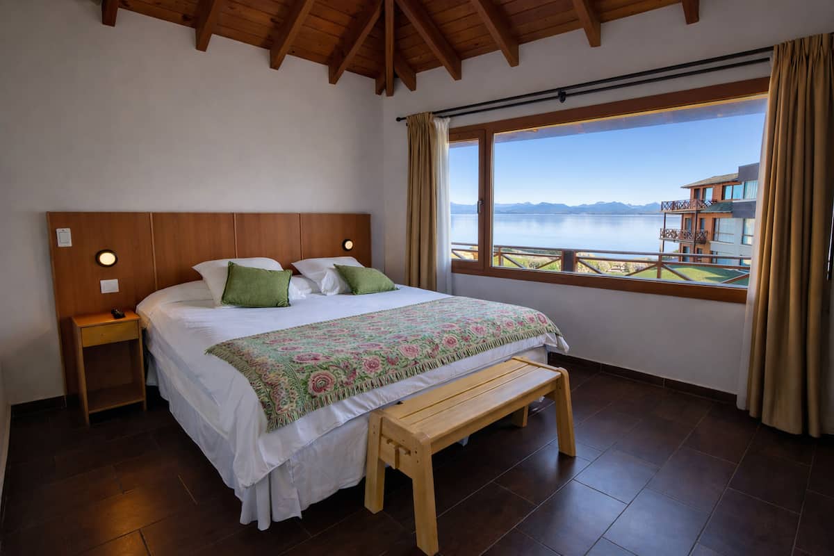Un dormitorio con cabecera de madera, cama con colcha floral, una banca, mesita de noche y ventana con vistas a un cuerpo de agua y edificios.