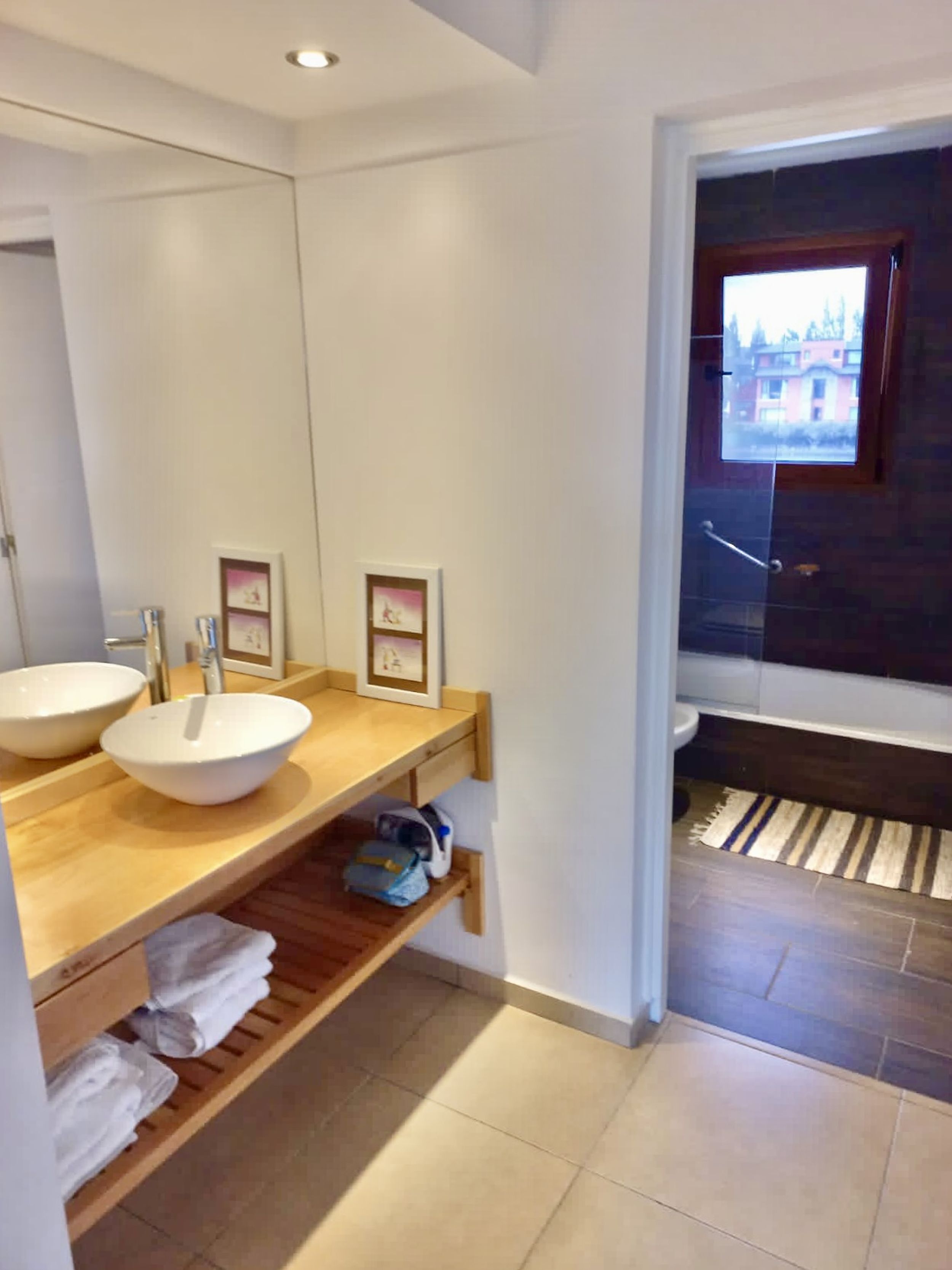 A modern bathroom with two large white sinks, a wooden vanity, and a large mirror. There are framed pictures on the wall, and a view of the outside through a window.