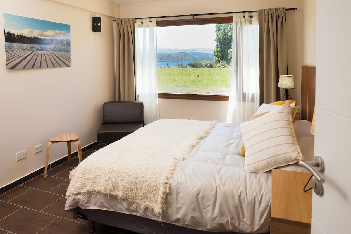 Un dormitorio con una cama, vistas a un lago, un taburete pequeño y una lámpara.
