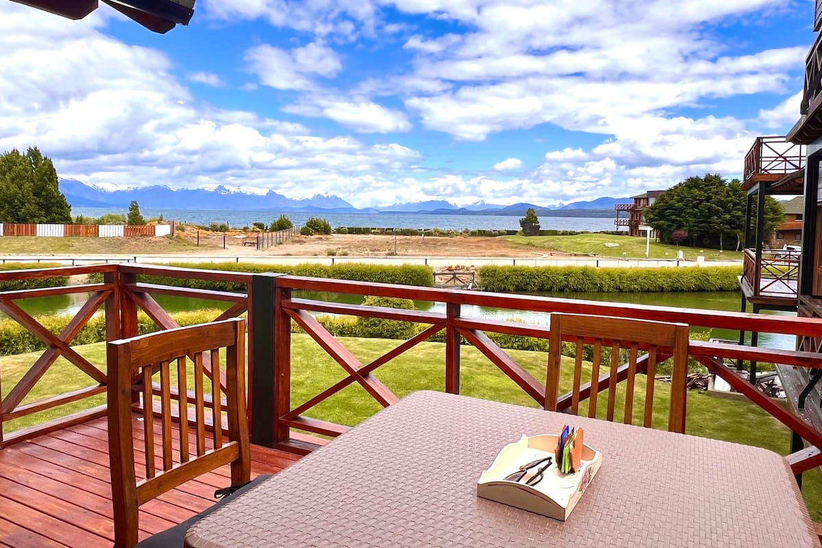 Una terraza de madera con una mesa y sillas, con vista a un lago y montañas.