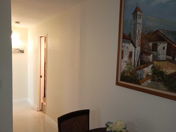 A dining area with a table and chairs, a painting of a church on the wall, and a hallway leading to another room. - Private accommodation Located 10 minutes from downtown, 5 from airport (West Palm Beach)