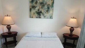 Une chambre à coucher avec un lit, deux tables de chevet avec des lampes, un tableau abstrait accroché au mur et un rideau à motifs.