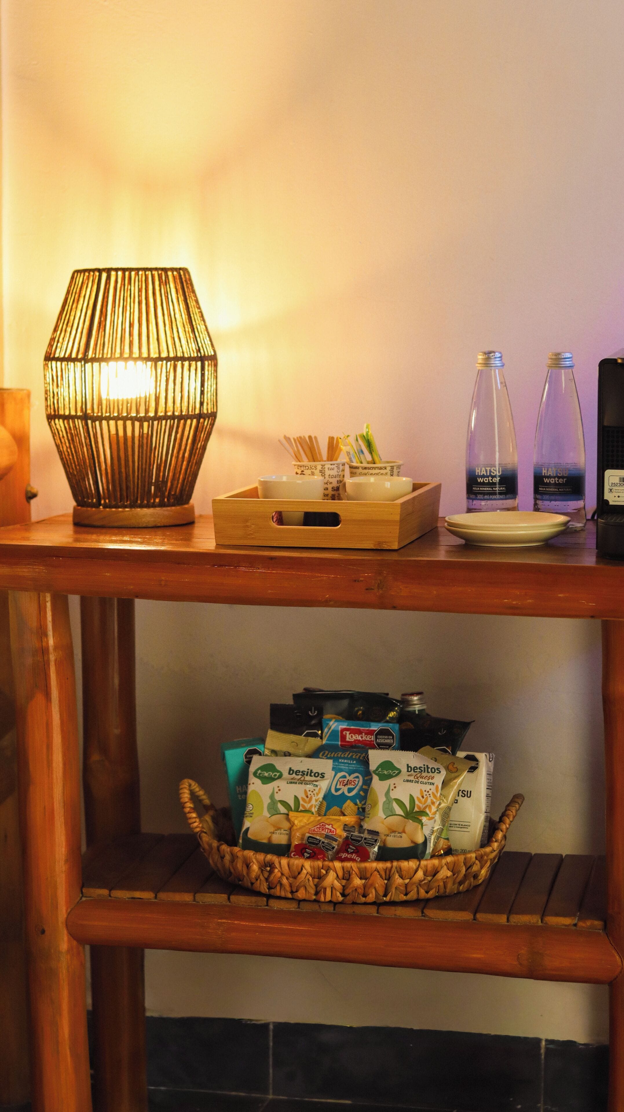 Una mesa de madera con una lámpara, dos botellas de agua, una bandeja con cuencos y una cesta de aperitivos.