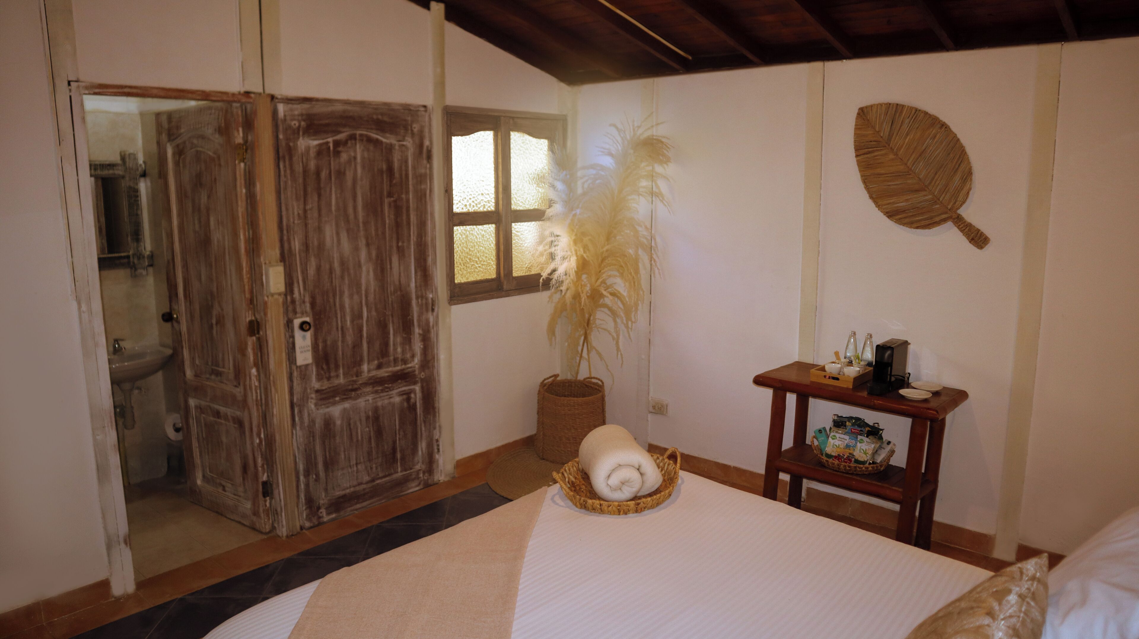 A room with a bed, a wooden door, a small table with a basket, a woven basket, and a wall-mounted decorative item.