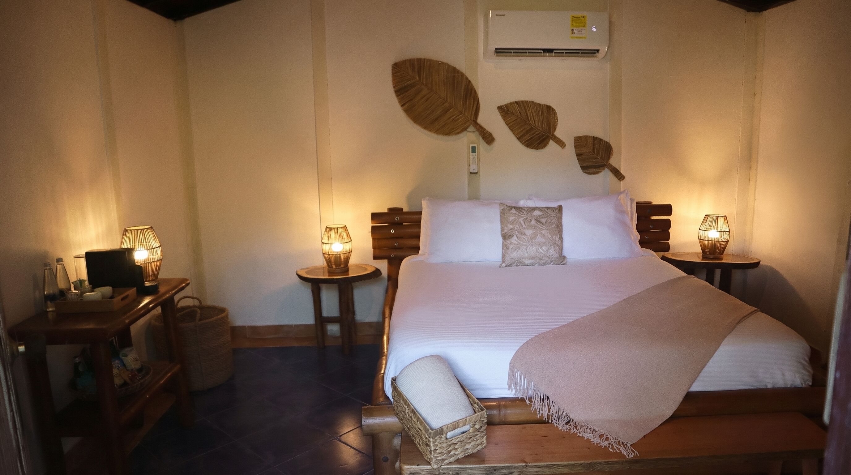 Un dormitorio ordenado con una cama grande, dos mesitas de noche con lámparas, una mesita auxiliar pequeña, una cesta de mimbre y un aire acondicionado de pared.