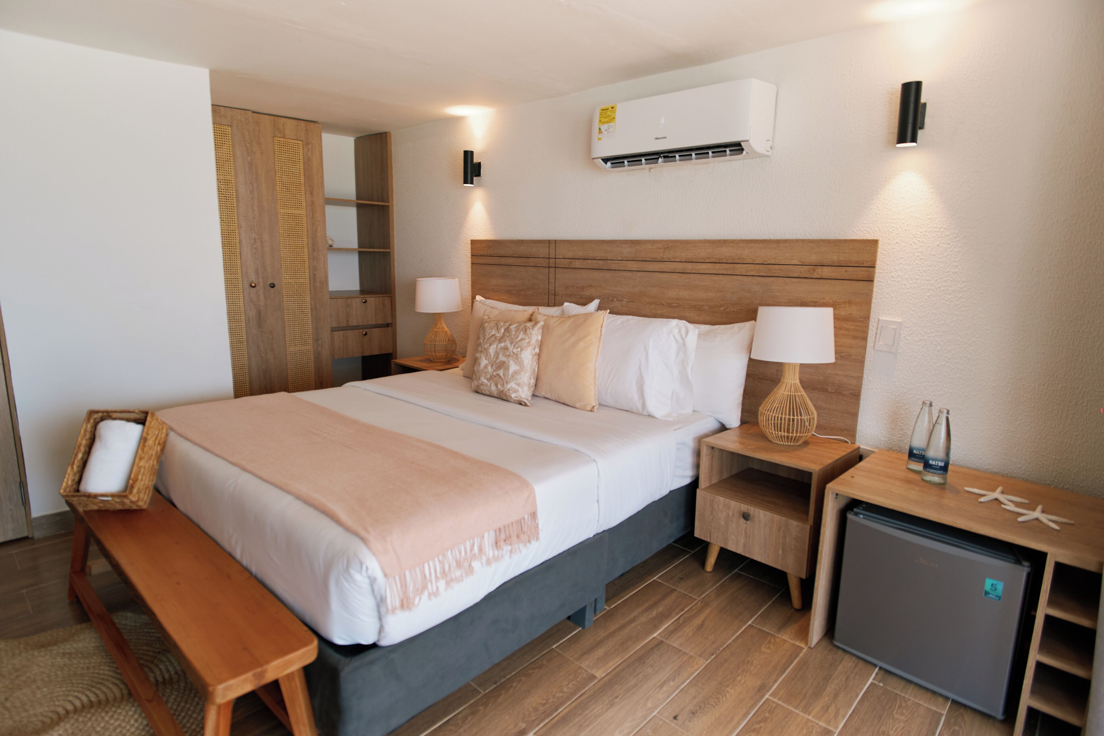 Een hotelkamer met een bed, nachtkastjes, een bankje, een kleine koelkast en een airconditioning.