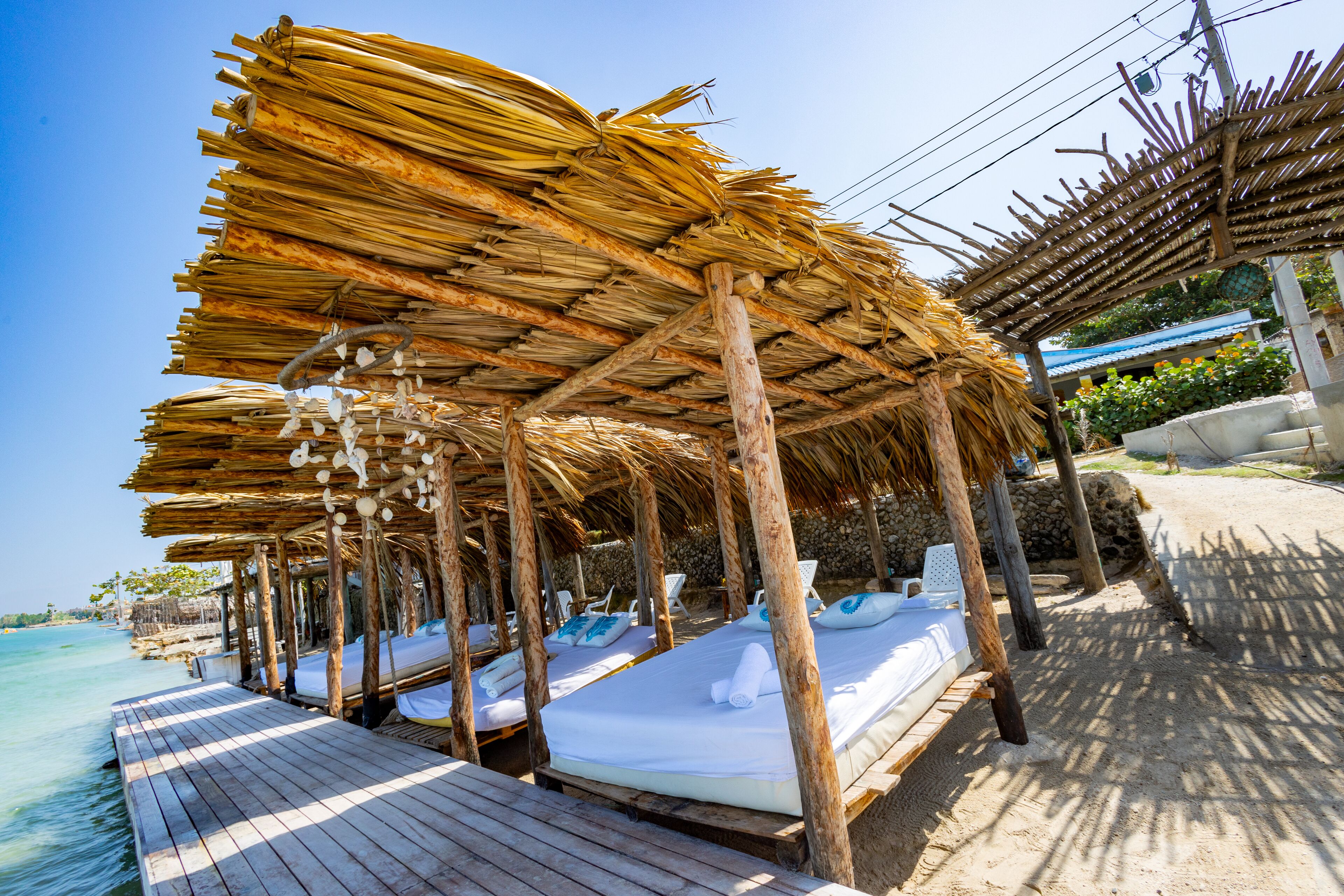 A beachside resort with thatched-roof cabanas, wooden deck, and white beds with blue pillows.