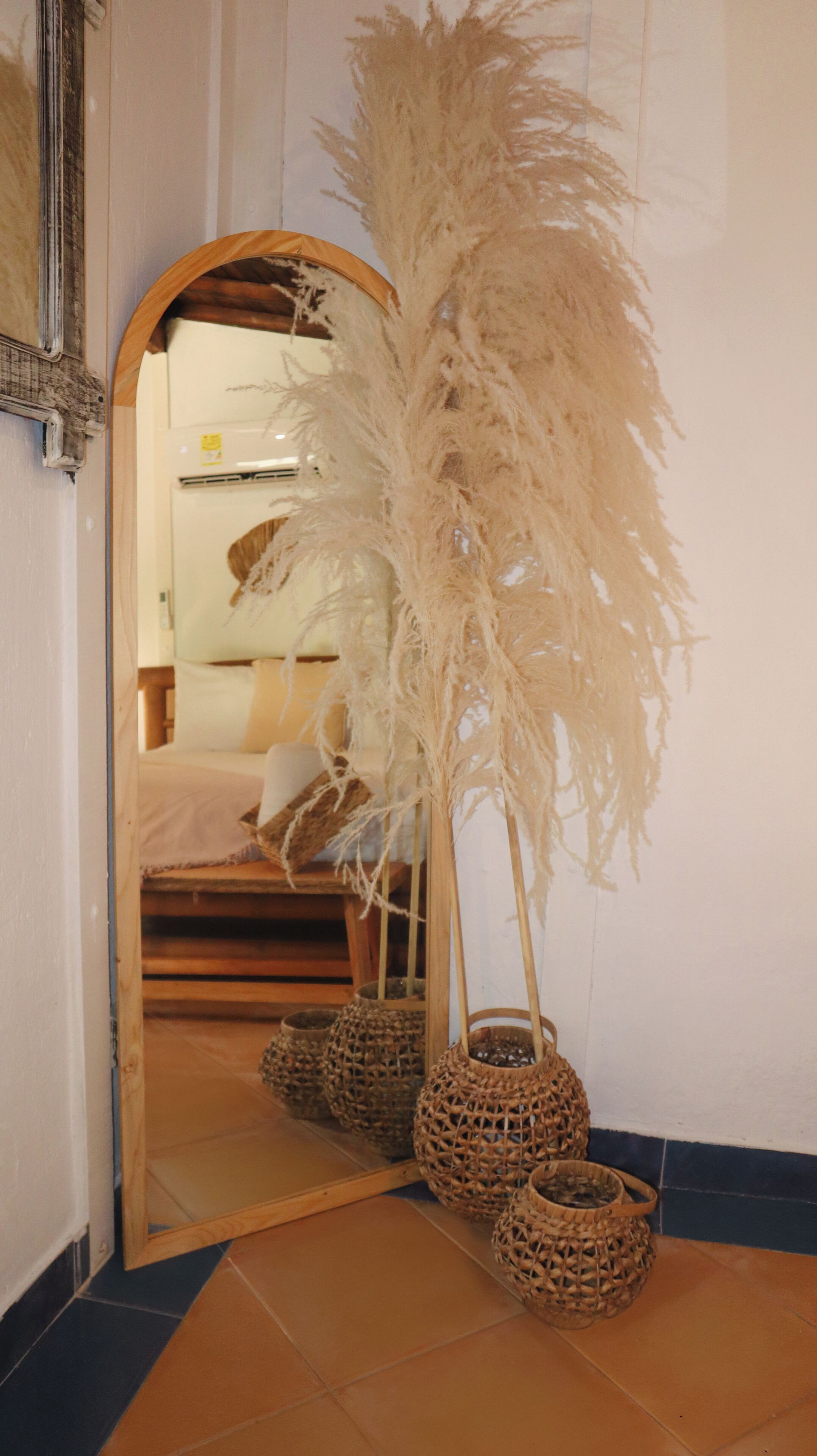 Um espelho reflete um quarto com uma cama, almofadas e uma mesinha de cabeceira. Em frente ao espelho, há três cestos trançados e uma planta decorativa alta.