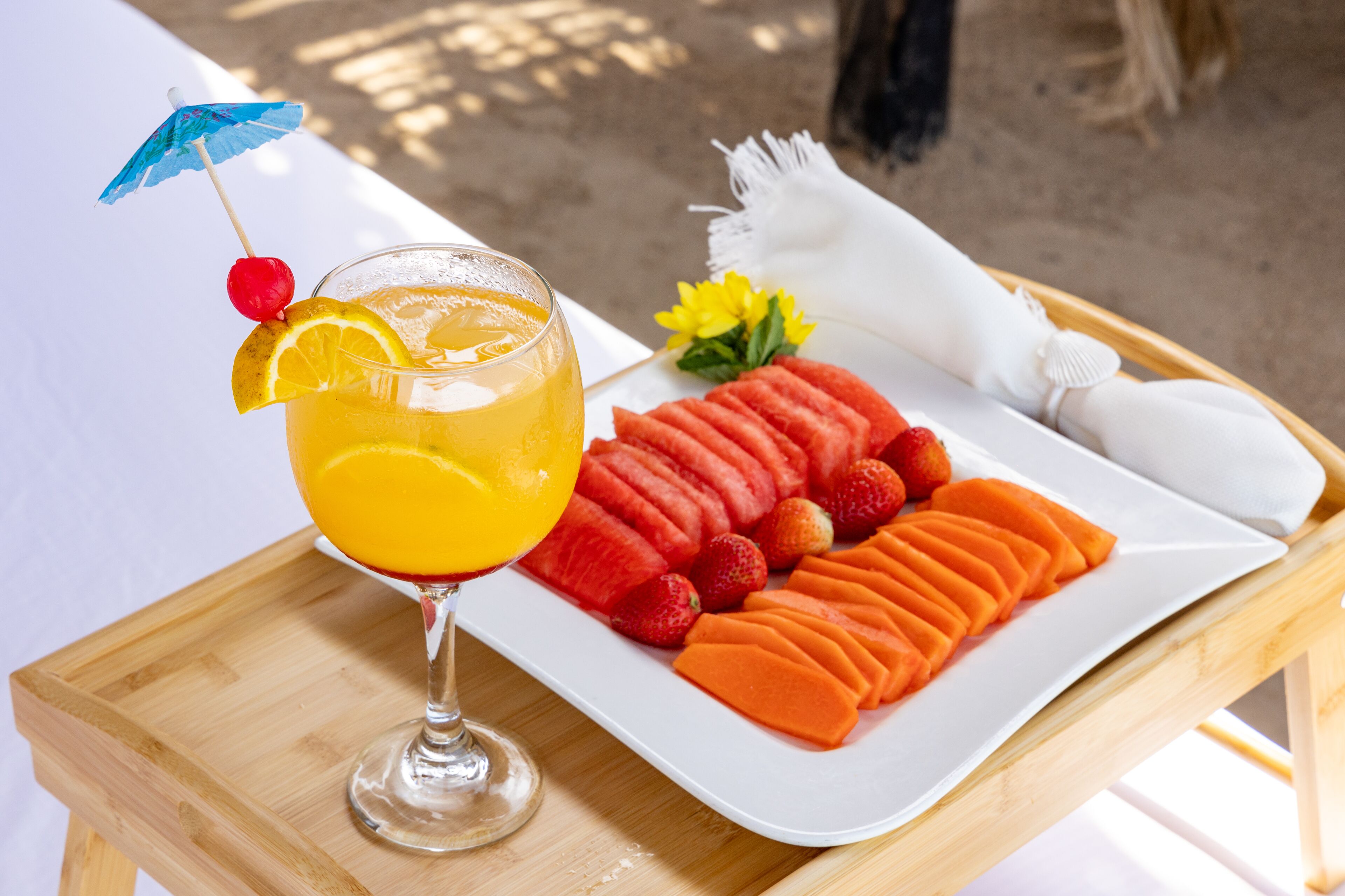 Una bandeja con un vaso de jugo de naranja, una rodaja de naranja, una cereza y una pajita, junto a un plato de sandía en rodajas, fresas y melón.