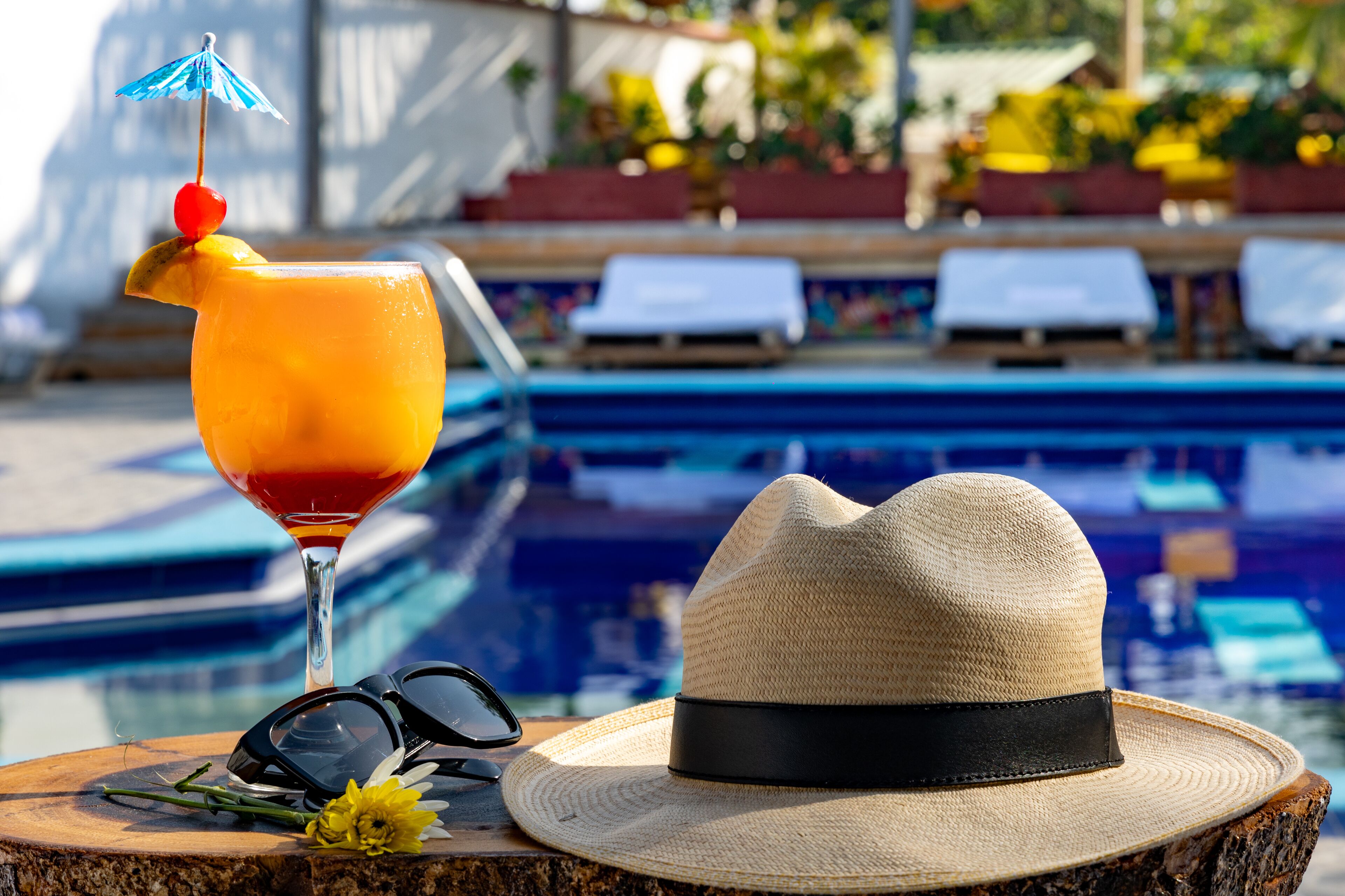 A poolside setting with a drink, sunglasses, a hat, and a flower on a wooden surface.