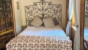 Ein Schlafzimmer mit Holzbalkendecke, einem verzierten Metall-Kopfteil, einer zierlichen Bettdecke mit Blumenmuster und einem großen Fenster mit Blick auf Bäume.