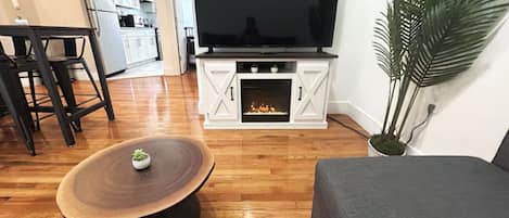 Un salón con una televisión de pantalla plana, una mesa redonda de centro, un sofá gris y una planta en maceta.