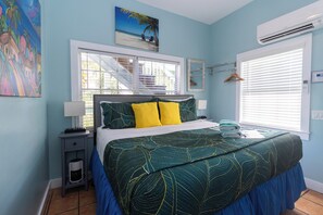 A bedroom with a bed, a nightstand, a window with blinds, a painting on the wall, and an air conditioning unit.