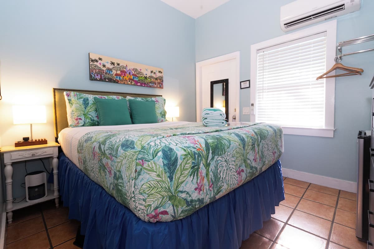 A bedroom with a bed, a nightstand, a lamp, a painting, a window with blinds, and a towel rack.
