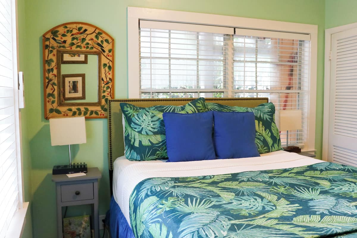 A bedroom with a bed, a nightstand, a mirror, and a window with blinds.