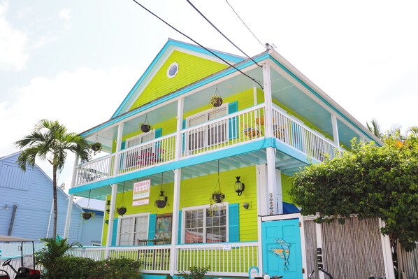 A two-story building with a bright green and blue exterior, a balcony on the upper floor, and hanging plants.