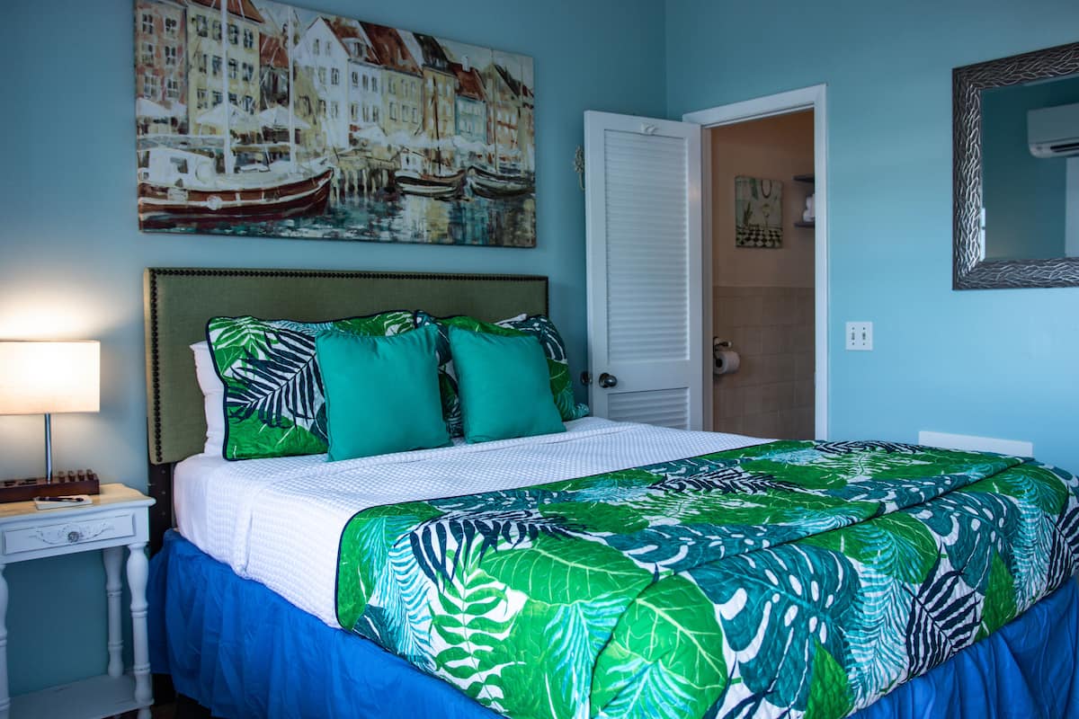 A bedroom with a bed, a nightstand, a painting of boats, a door, and a mirror.