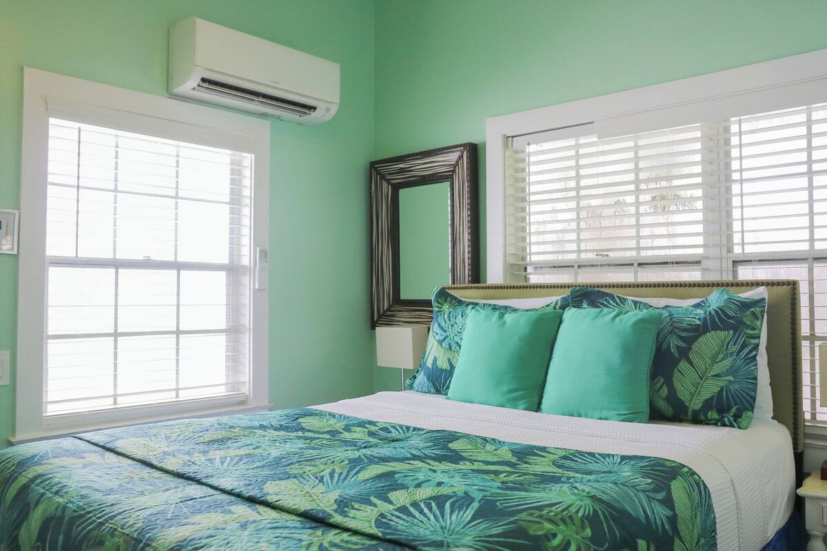 A bedroom with a bed, two windows with blinds, an air conditioning unit, and a mirror on the wall.