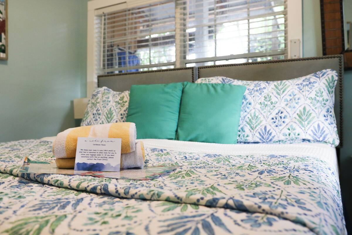 A neatly made bed with a floral bedspread, two green pillows, and a rolled towel.