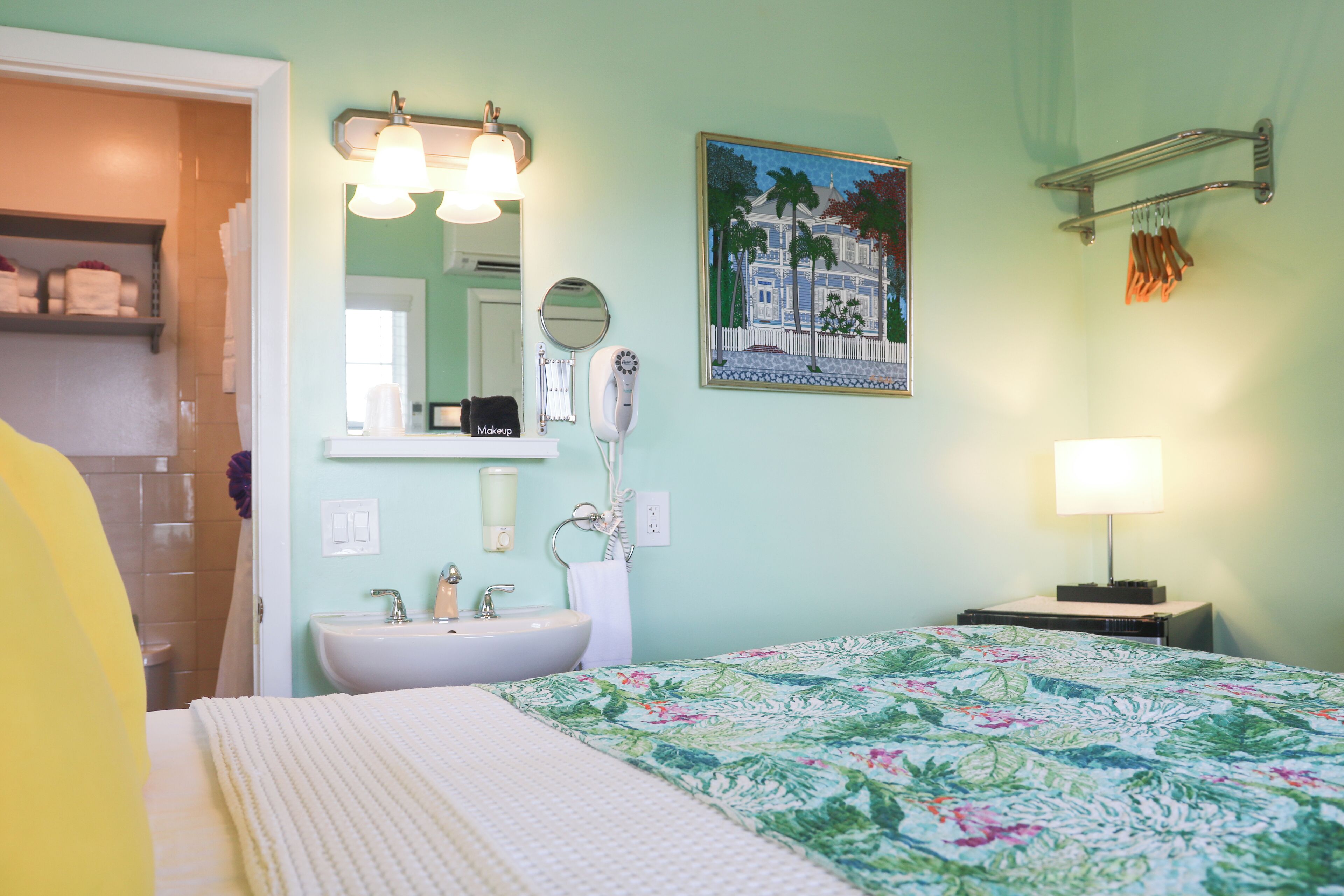 A bedroom with a bed, a bedside table, a lamp, a mirror, a sink, and a painting on the wall.