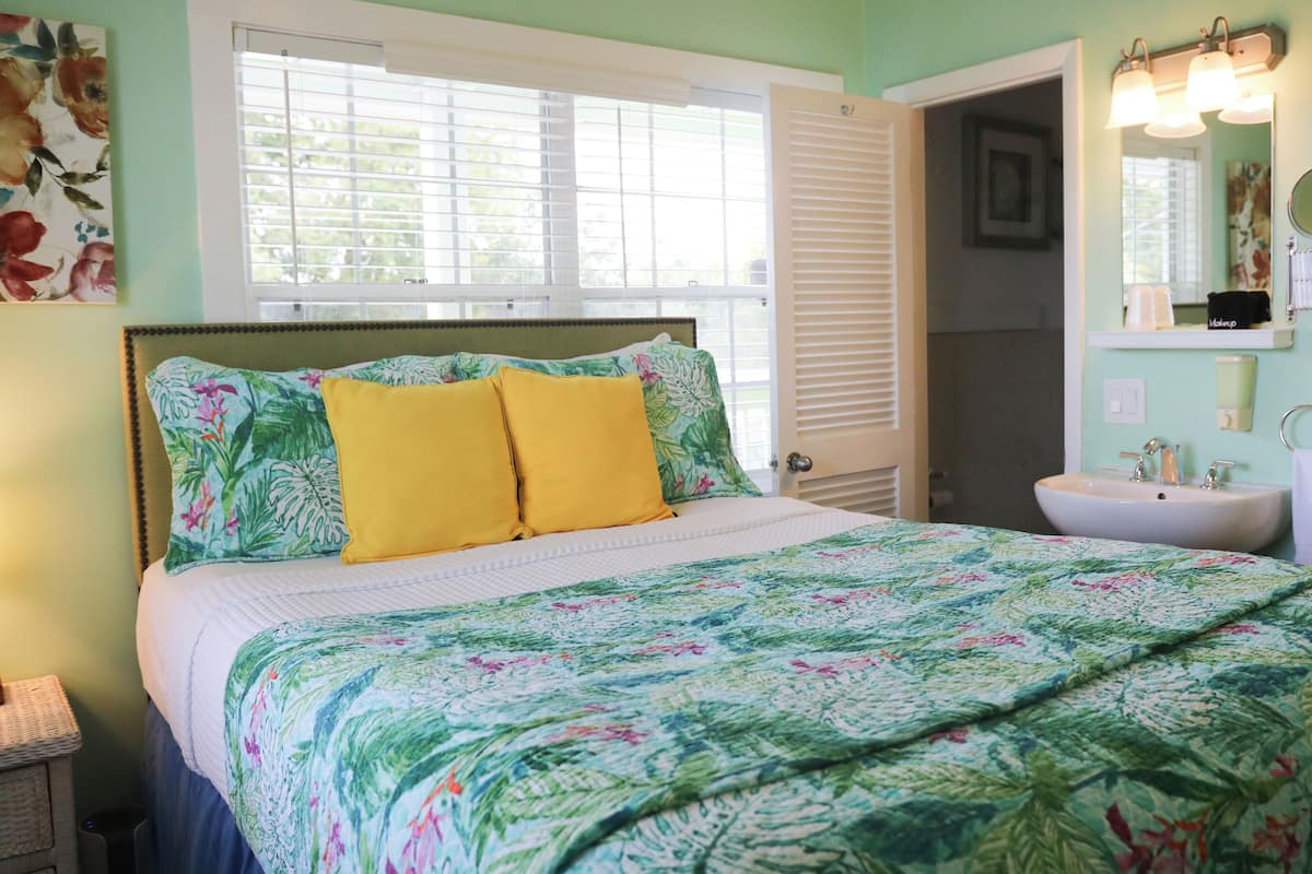 A bedroom with a bed, a window with blinds, a bedside table, and a bathroom with a sink and mirror.