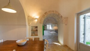 Un coin repas avec une table et des chaises en bois, un bol blanc, un luminaire suspendu et une arche en pierre menant à la cuisine.