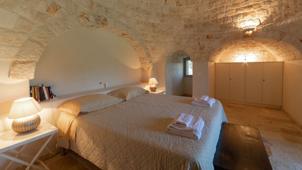 Une chambre confortable avec un plafond en pierre, un lit recouvert d’une couverture matelassée, une lampe de chevet et une petite table avec une lampe.