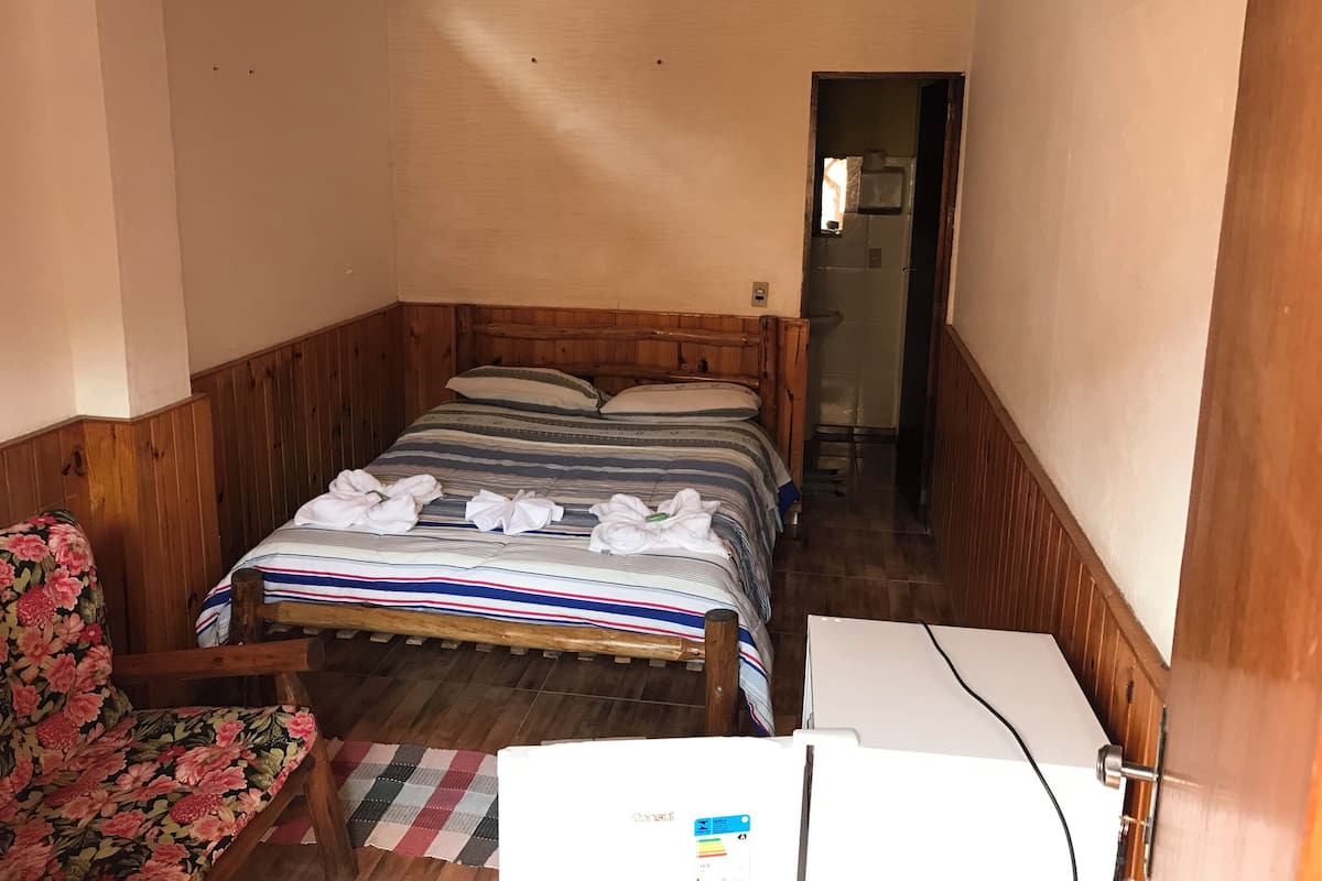 A small, cozy room with a wooden bed, a chair with floral upholstery, and a white refrigerator.