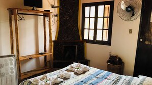 A room with a bed, a stone fireplace, a wooden shelf, a fan, and a window with shutters. - Pousada Cantinho da Montanha (Itatiaia)