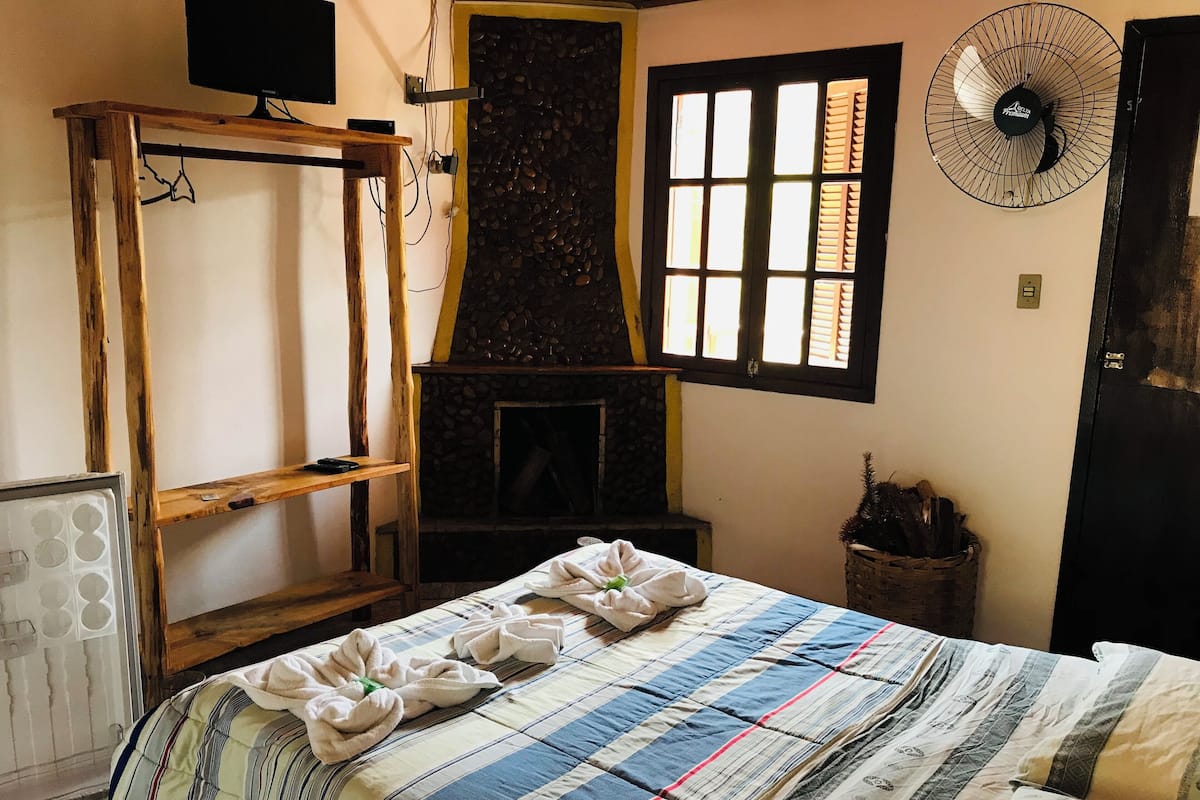 A room with a bed, a stone fireplace, a wooden shelf, a fan, and a window with shutters.