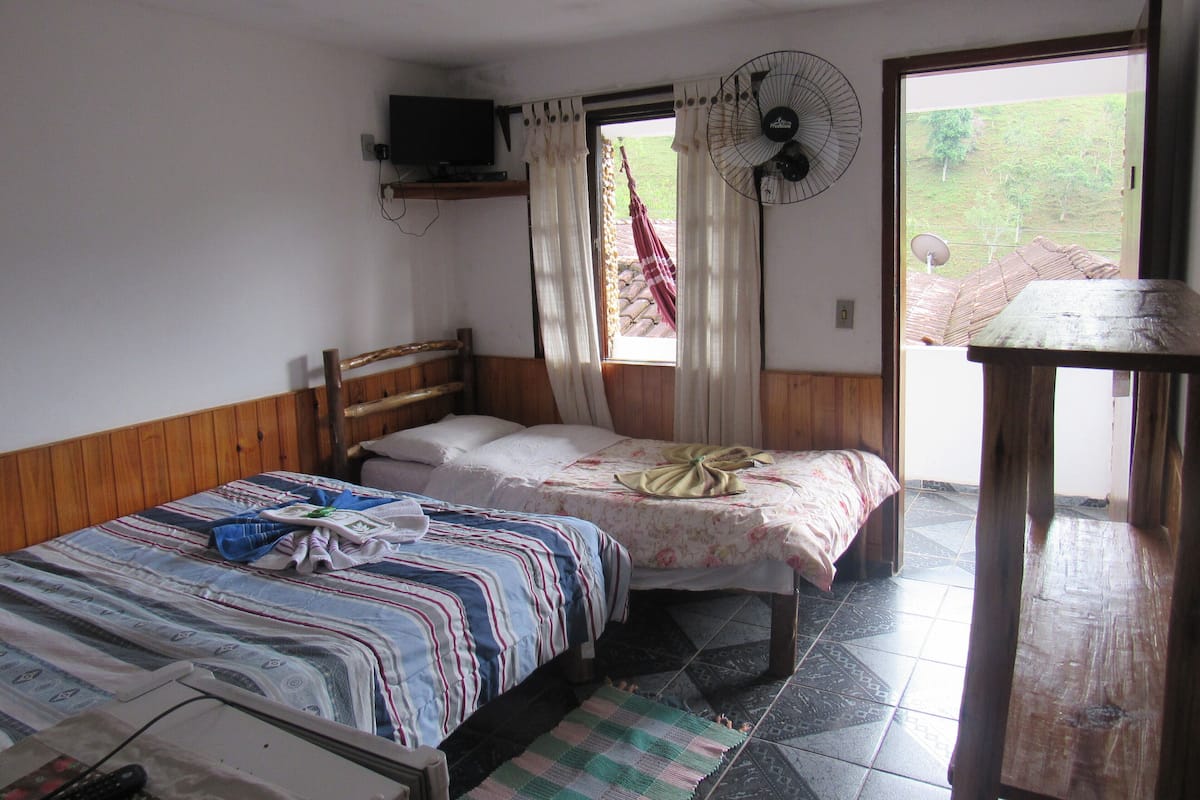 A bedroom with a wooden bed, a television on the wall, a window with curtains, and a fan.