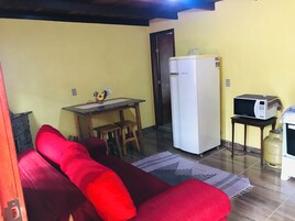 Un espacio habitable pequeño con un sofá rojo, una mesita de madera con un florero, un refrigerador blanco, un microondas sobre una mesita y un cilindro de gas.