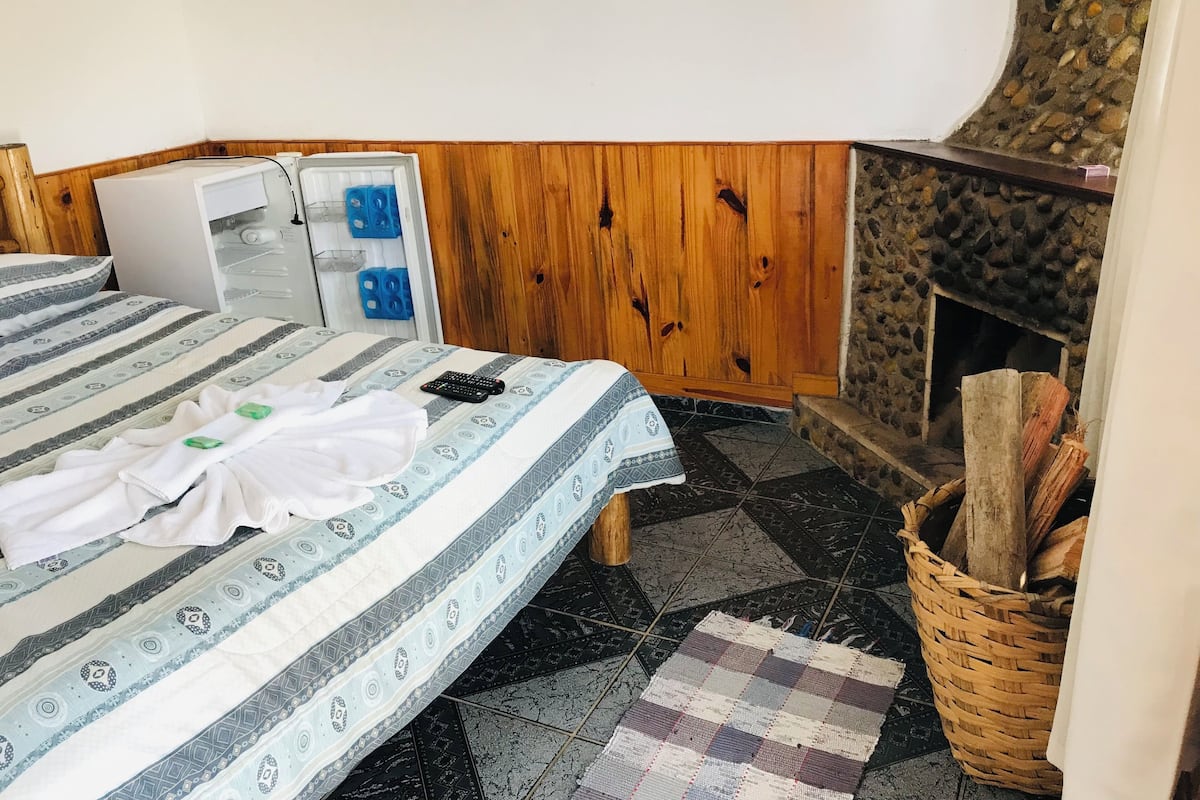 A bedroom with a bed, a small refrigerator, a stone fireplace, and a wooden wall.