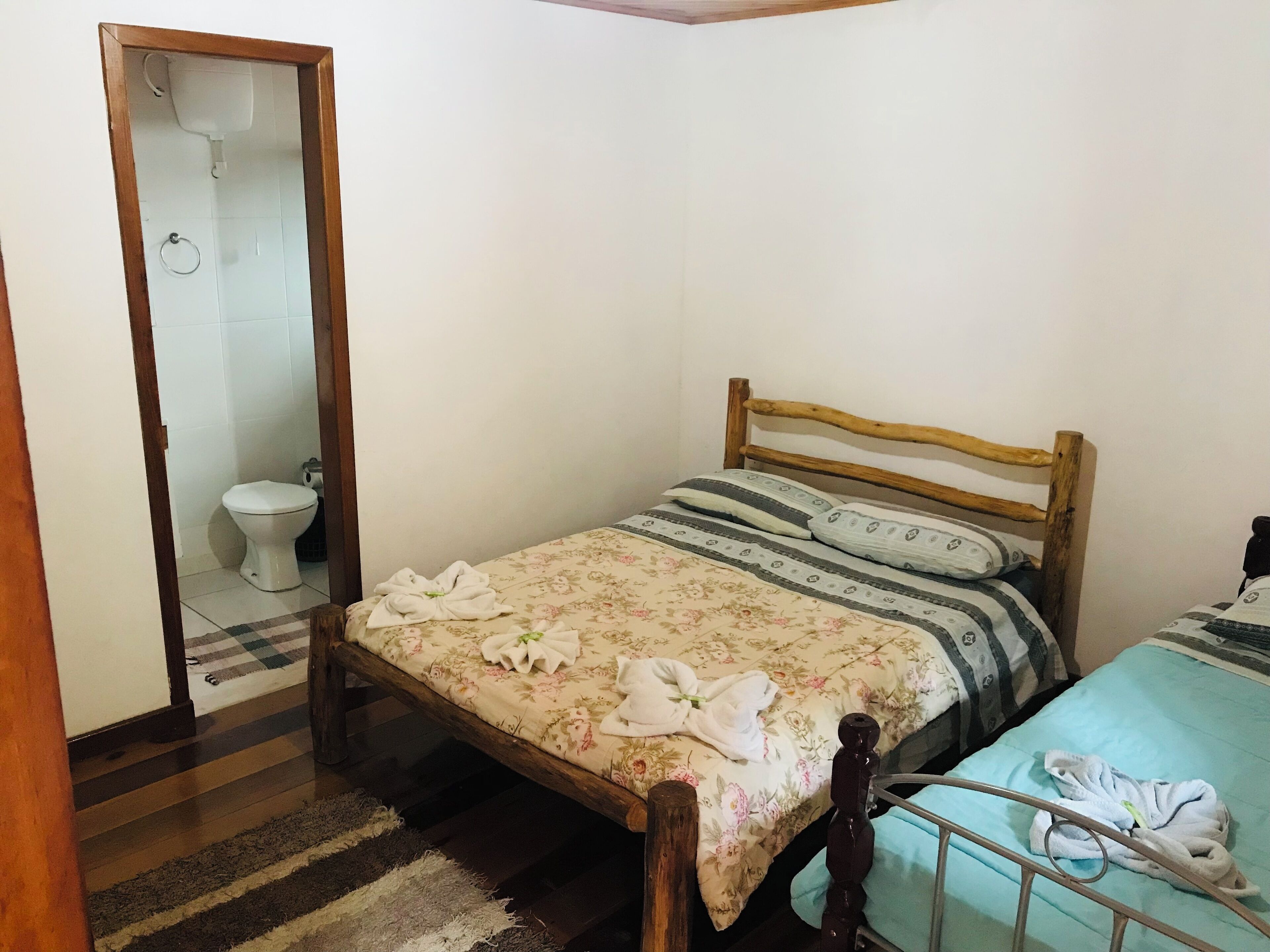 A bedroom with a wooden bed, a small bedside table, a single bed with a floral bedspread, a blue bedspread, and a white toilet.