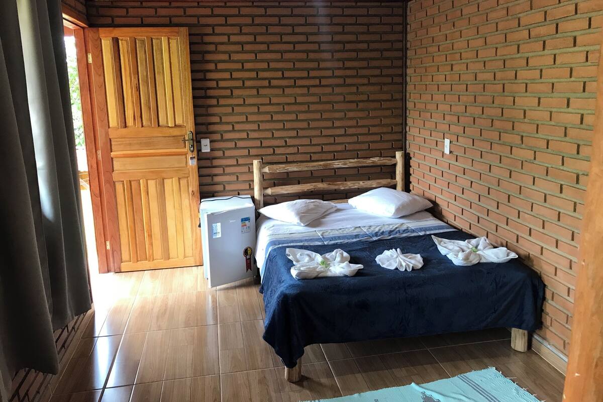 A room with a bed, a wooden door, a small white appliance, and a brick wall.