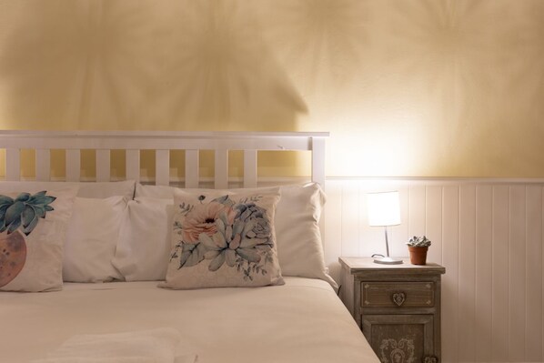Un dormitorio con una cama blanca, almohadas estampadas, una mesita de noche con lámpara y un mueble auxiliar con una planta.