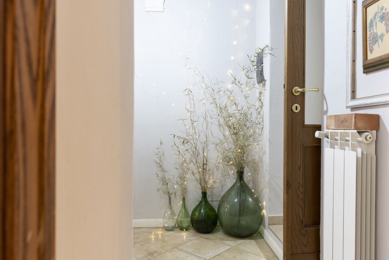 Un couloir avec une porte en bois, un radiateur et trois vases en verre vert contenant des plantes séchées et des guirlandes lumineuses.