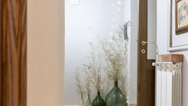 Un pasillo con una puerta de madera, un radiador y tres jarrones de vidrio verde con plantas secas y luces de hadas.