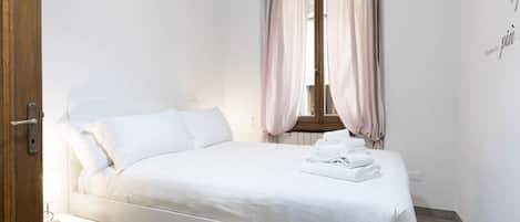 A neatly made bed with white linens, a bedside table with a lamp, and a window with pink curtains.