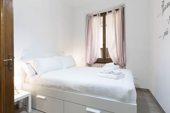 A neatly made bed with white linens, a bedside table with a lamp, and a window with pink curtains.