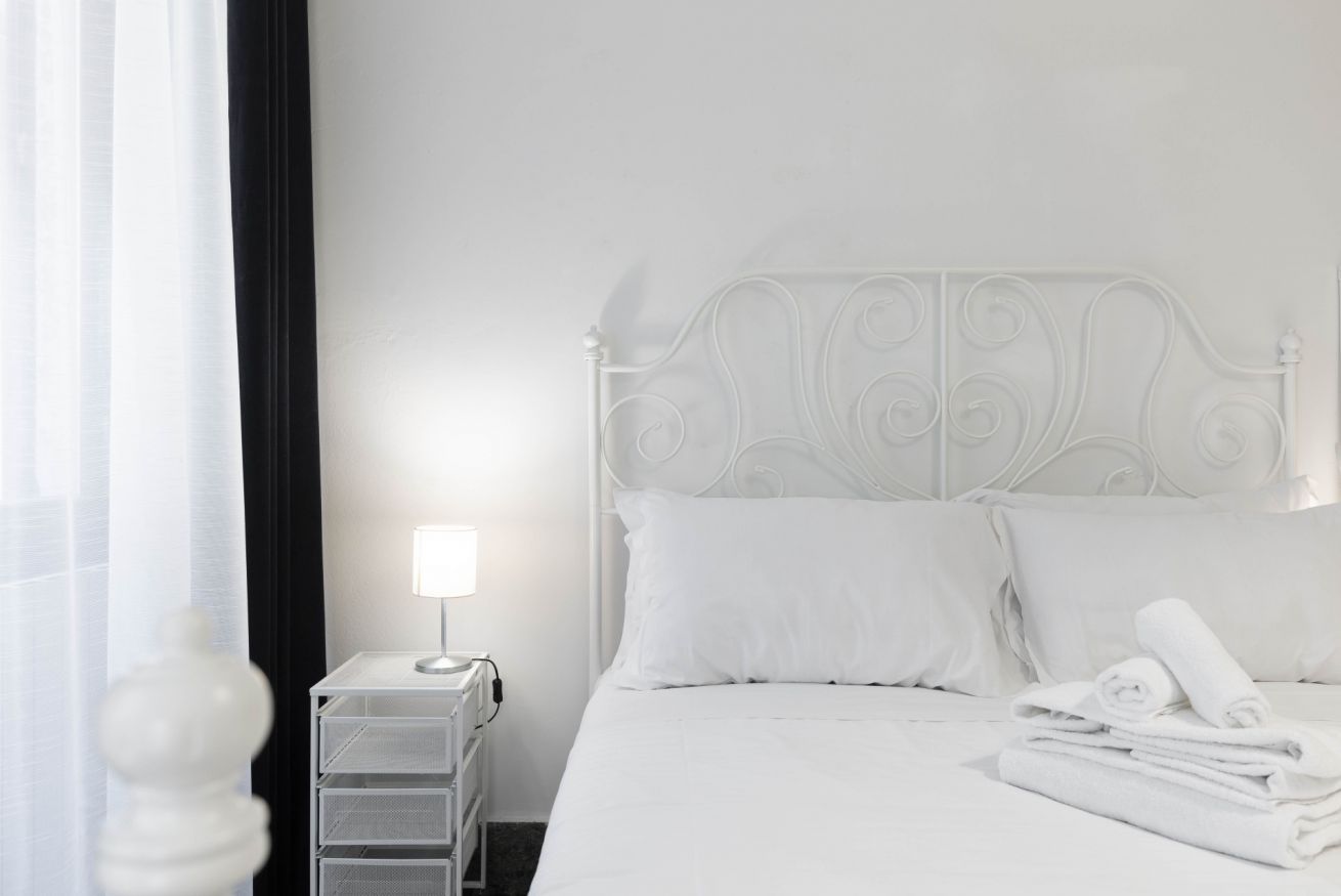 A white bed with a metal headboard, a bedside table with a lamp, and rolled towels on the bed.