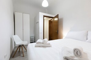 A hotel room with a bed, a white chair, a wardrobe, and a door leading to another room.