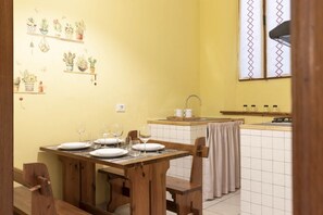 A dining area with a wooden table set for four, a kitchen with a tiled backsplash, and a window with patterned curtains.