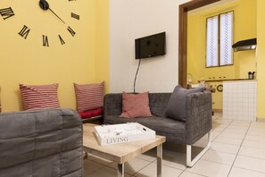 A living room with a sofa, a coffee table with a tray labeled "LIVING", a wall clock, and a kitchen area in the background.