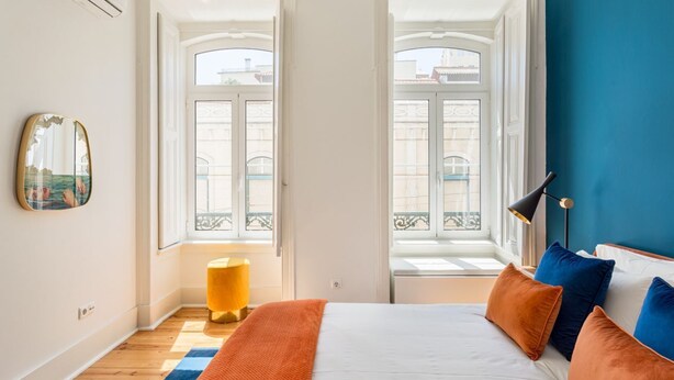 Una camera da letto con un letto, due finestre, uno specchio e uno sgabello giallo.