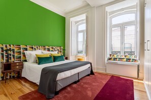 A bedroom with a green wall, a bed with multiple pillows, a window seat with colorful cushions, and a wooden floor. - JOIVY Exquisite apt w/ patio in Graça (Lisboa)