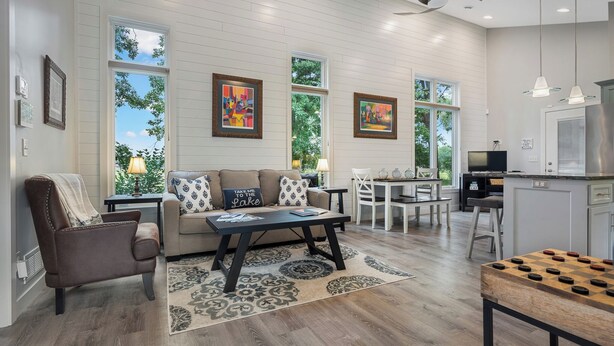 A spacious living area with a sofa, coffee table, and a chess table. There are large windows with artwork, a kitchen area, and a dining table.