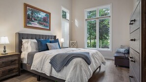 A bedroom with a large bed, a nightstand, a dresser, and a window with a view of trees.